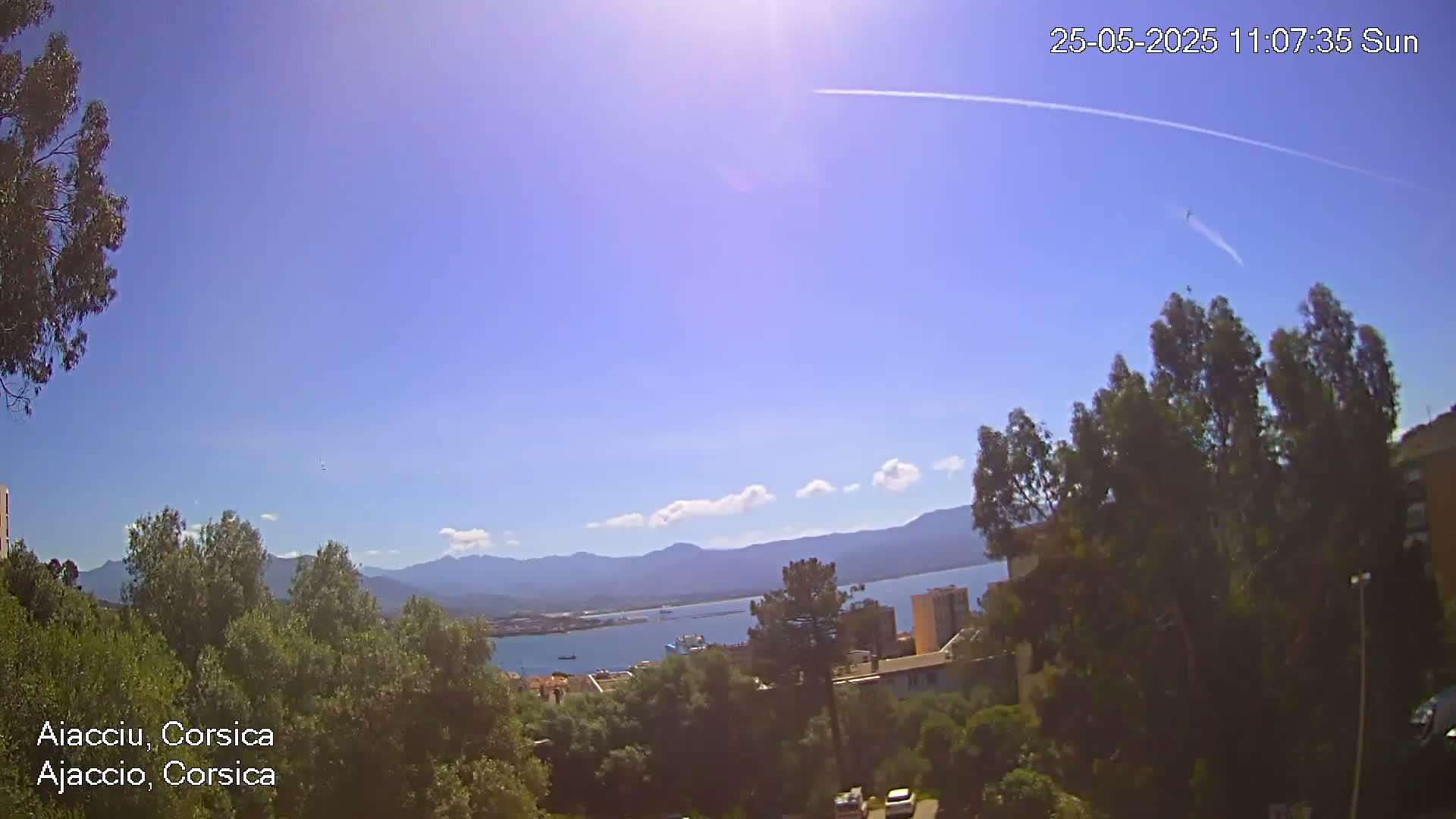 A sunny, mostly clear day reveals a view of a coastal town nestled among green trees and hills, with a calm body of water and distant mountains visible in the background.