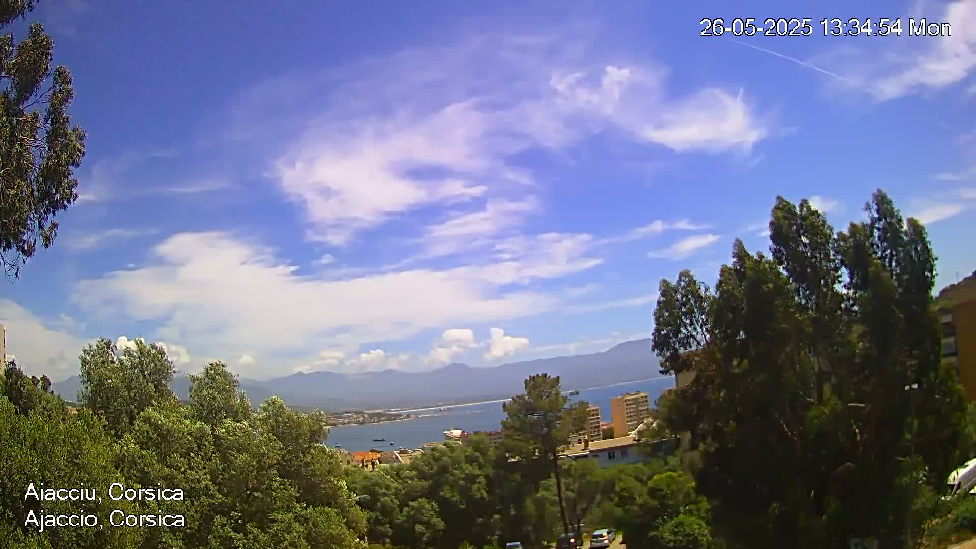 A partly cloudy blue sky overlooks a coastal city with green trees and mountains in the distance.