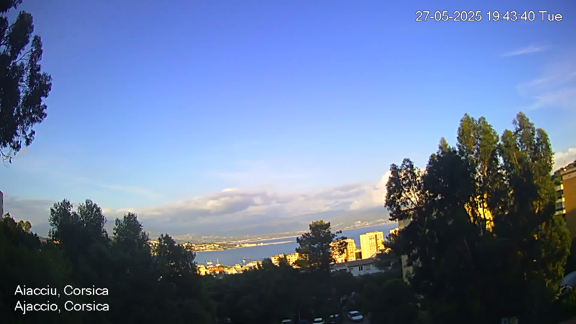 A mostly clear, blue sky with a few scattered clouds overlooks a coastal city partially obscured by trees.