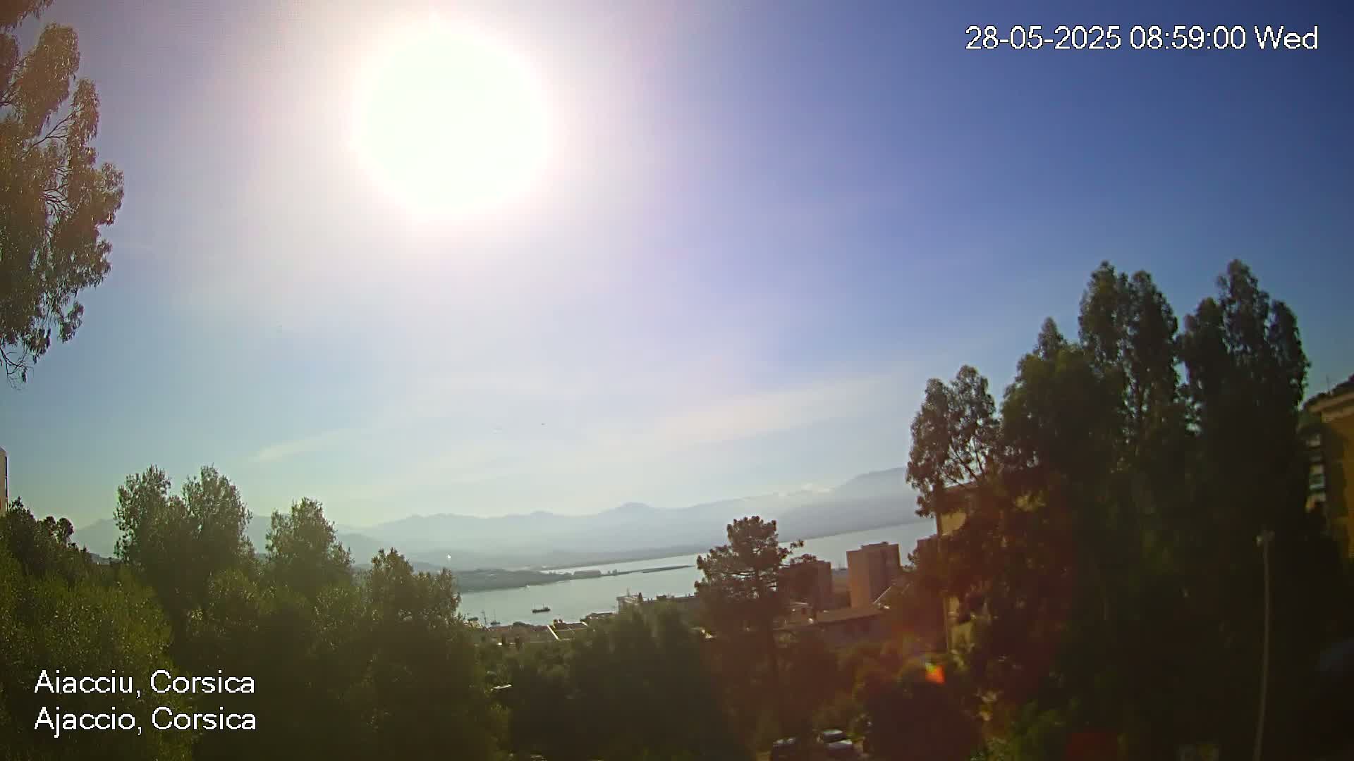 A sunny, hazy day reveals a coastal view of a city nestled among green hills and a calm body of water, with distant mountains visible in the background.