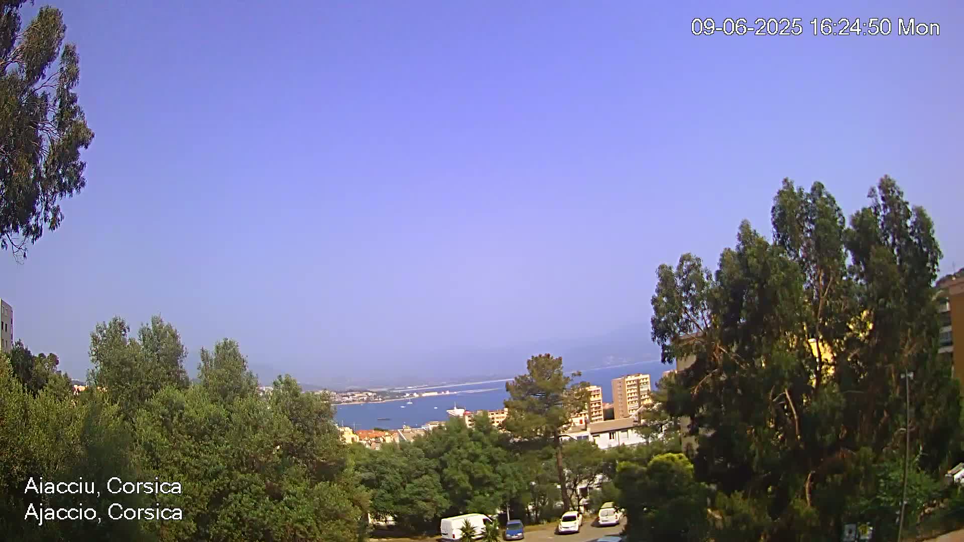 A clear, sunny day overlooking a coastal city with green trees and a few parked cars.