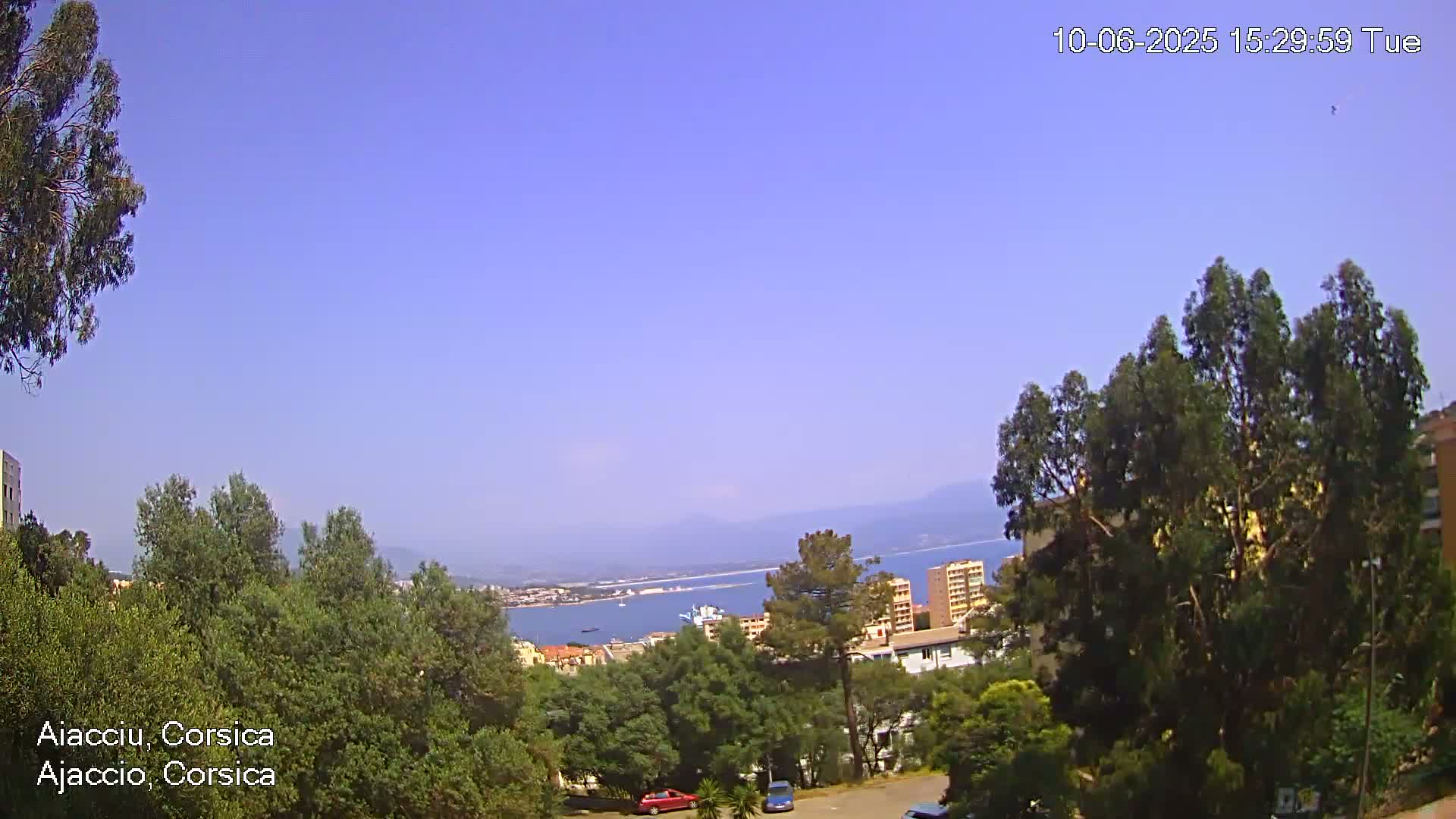 A clear, sunny day overlooks a coastal town nestled among green trees and buildings, with a body of water and distant mountains visible in the background.
