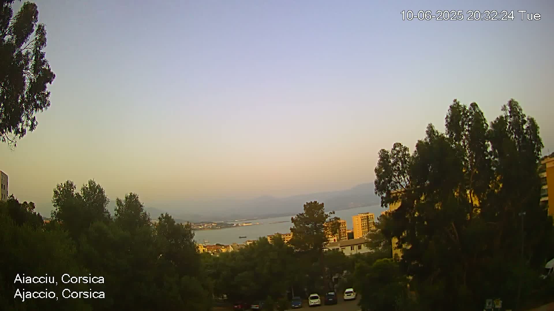 An evening view of a coastal town nestled among trees, with a calm body of water and distant mountains visible under a hazy, light-colored sky.
