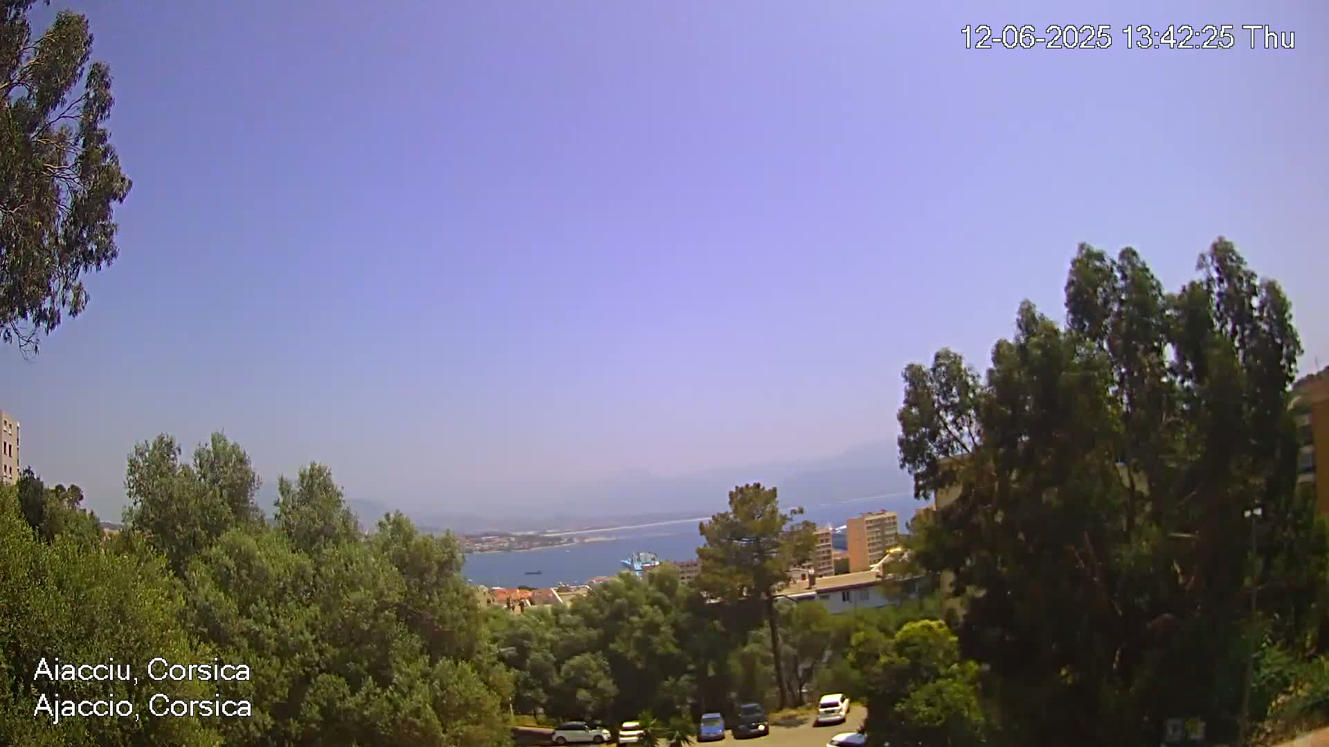 A clear, sunny day overlooks a coastal city nestled among green trees and a calm blue sea.