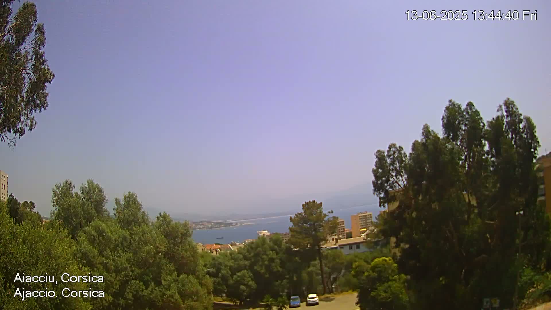 A clear, sunny view overlooks a coastal town nestled among green trees and hills, with a calm sea visible in the distance.