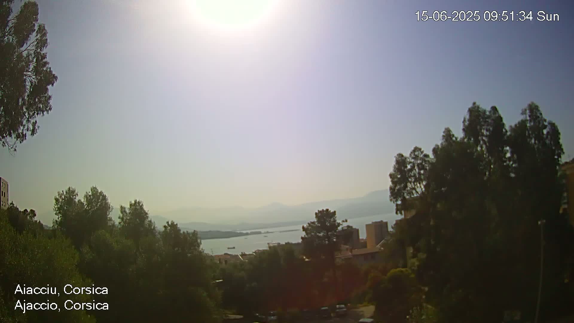 A hazy, sunny day overlooks a calm body of water, with green hills, trees, and buildings in the foreground.