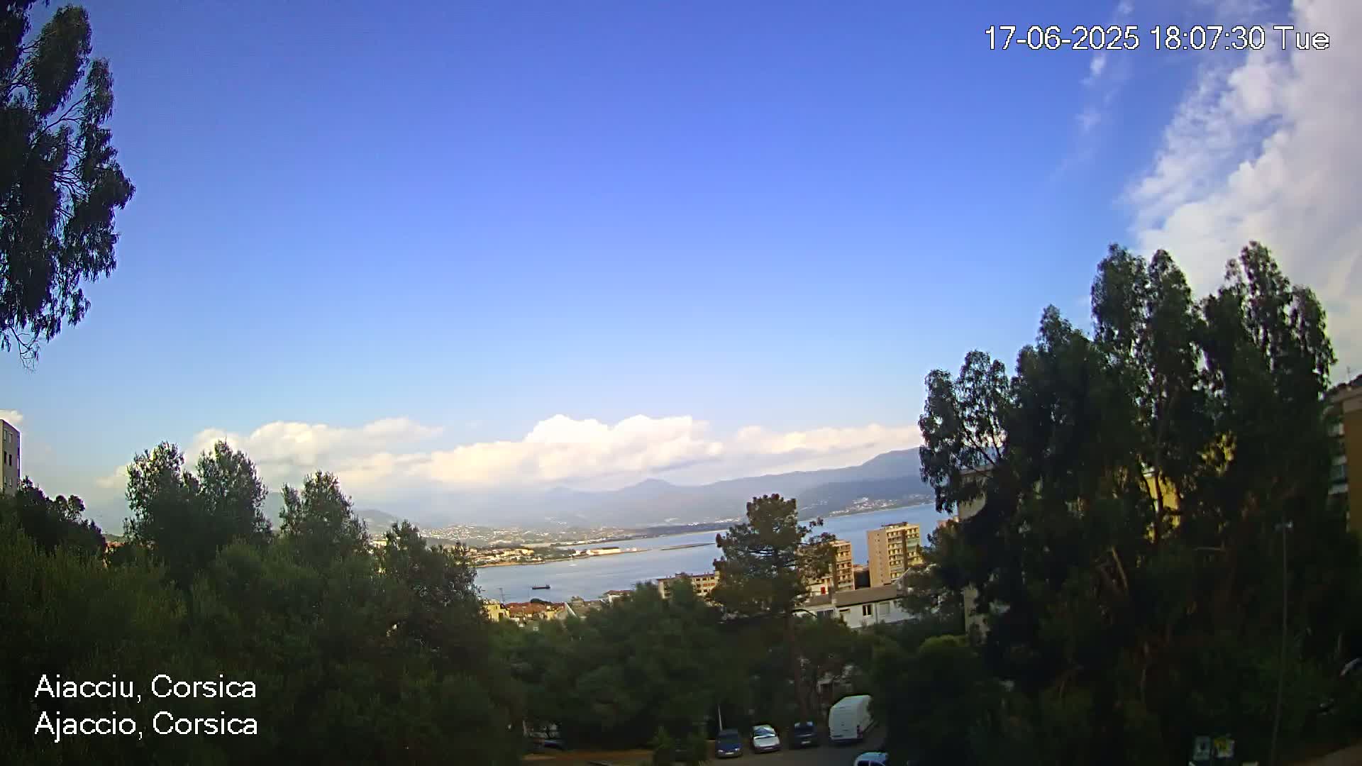 A partly cloudy, blue sky overlooks a coastal city with green trees and a body of water, mountains in the distance.