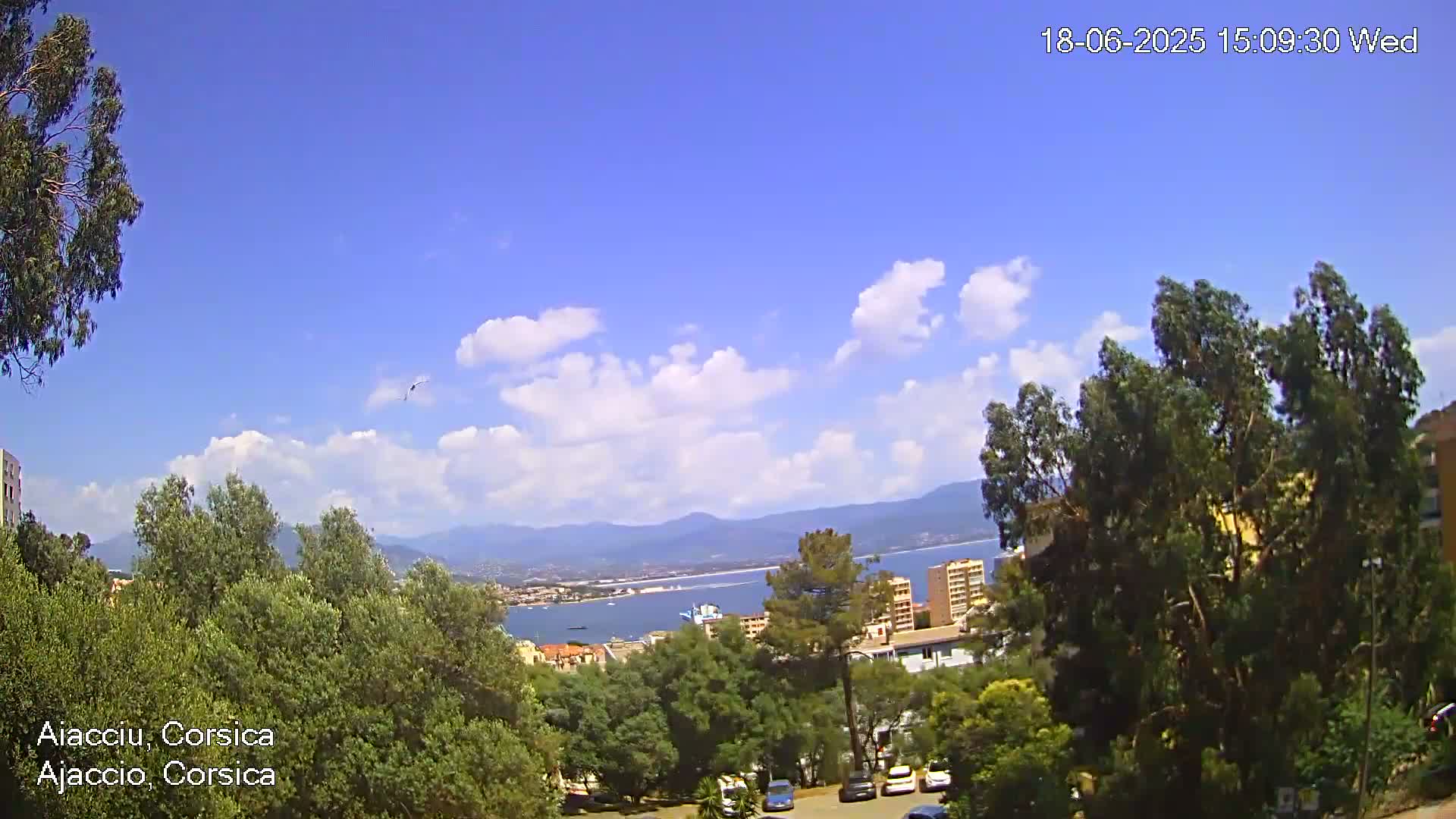 A partly sunny day overlooking a coastal town with a body of water and mountains in the background, viewed from a hilltop amidst green trees.