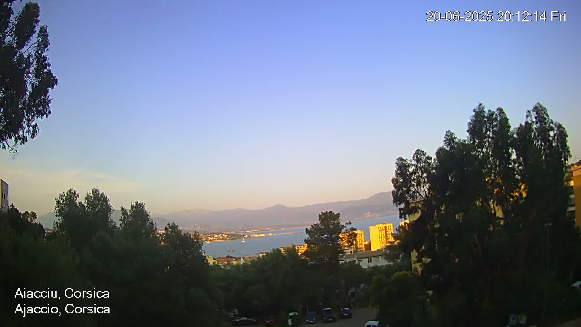 A clear evening sky over a body of water, backed by mountains and silhouetted trees and buildings.