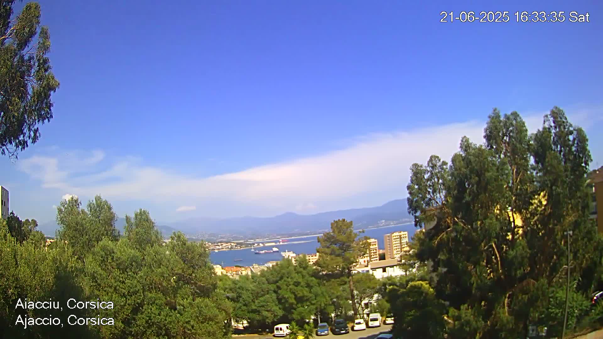 A partly cloudy, sunny day overlooks a coastal city with a harbor and mountains in the distance, as seen from a hilltop vantage point amongst lush green trees and parked cars.