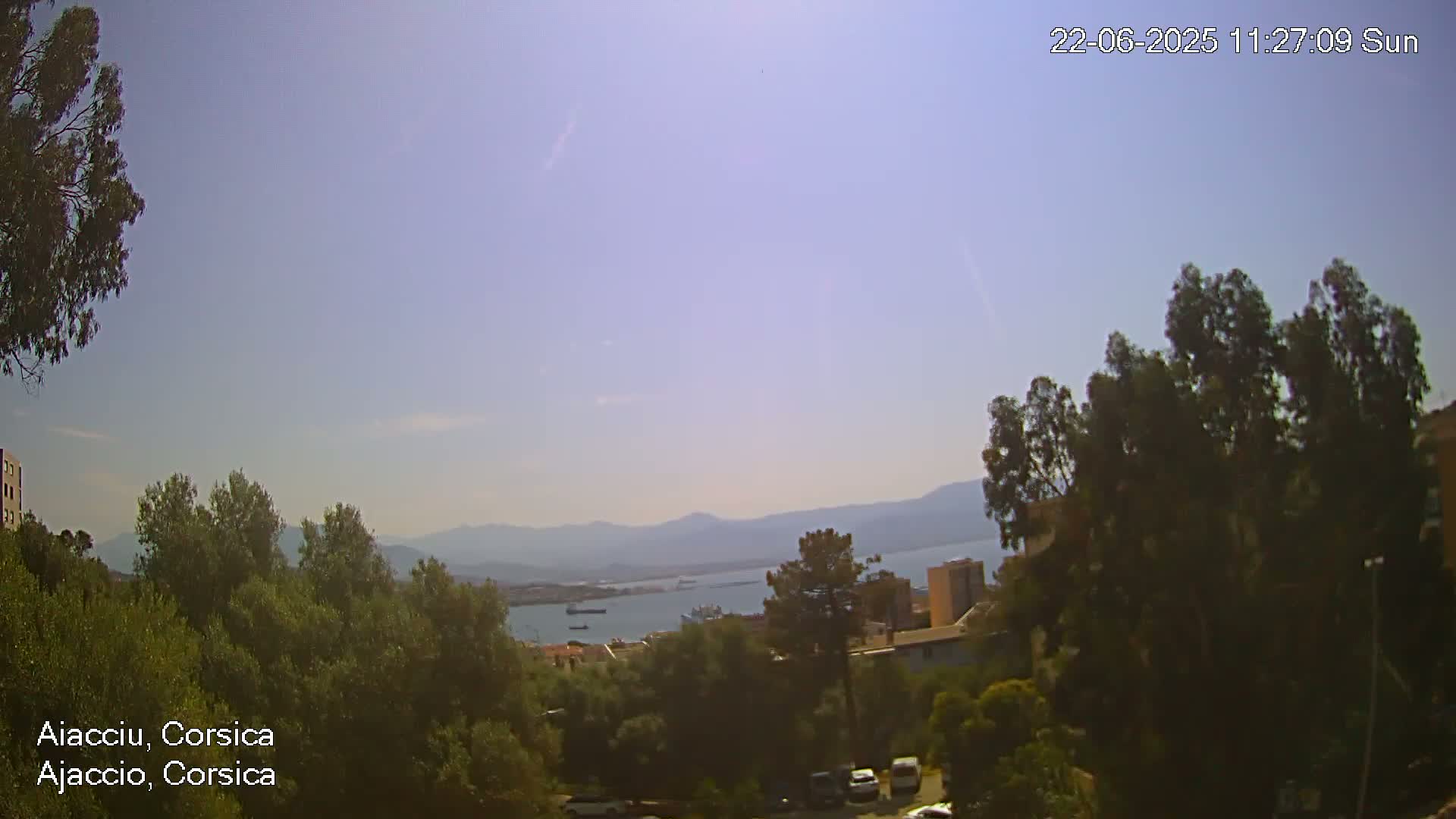 A sunny day reveals a view of a body of water with mountains in the distance, observed from an elevated vantage point amongst green trees and buildings.