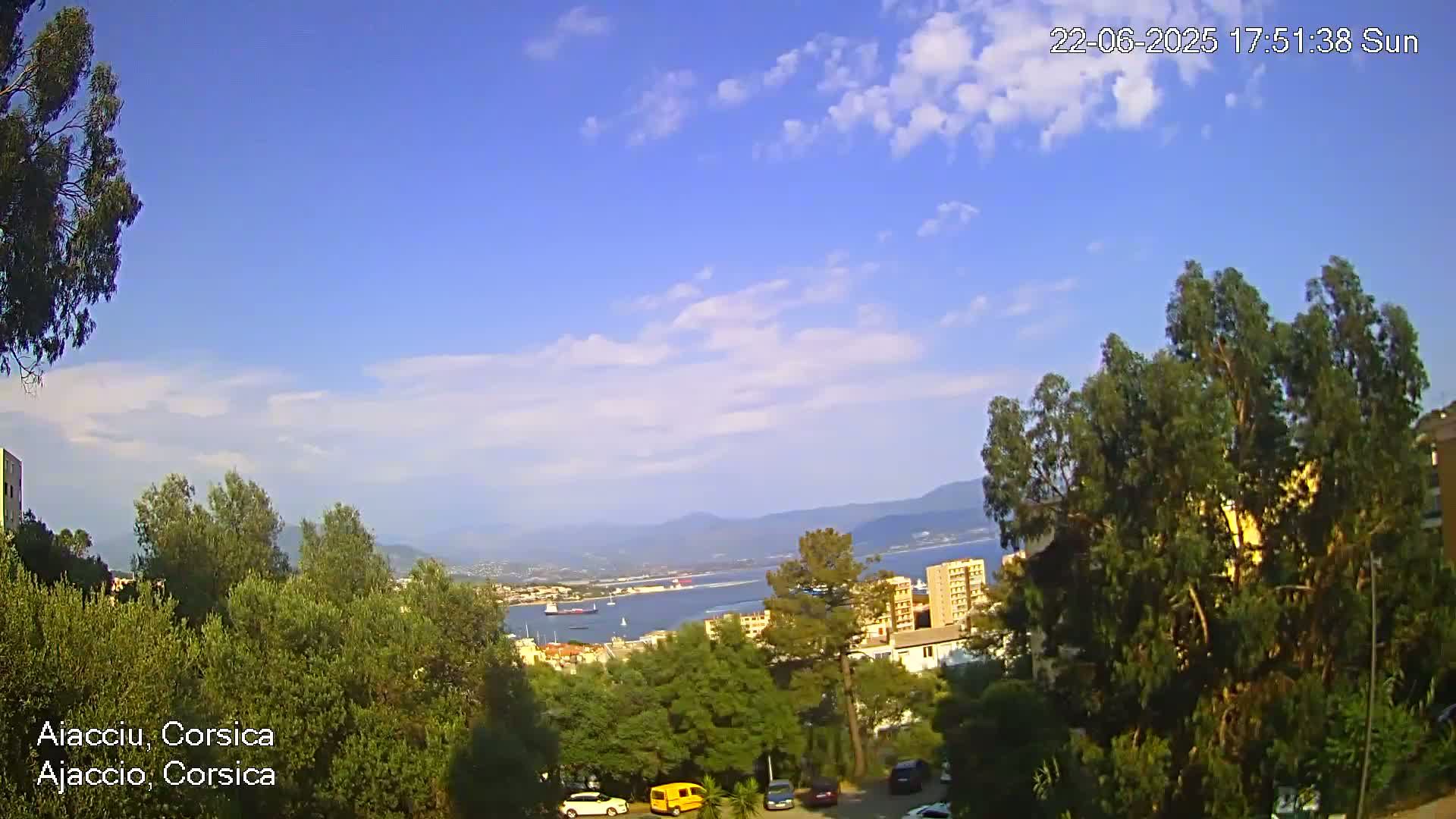 A sunny day with a few clouds reveals a coastal view of a city nestled among green trees and hills, a body of water with vessels visible in the distance.