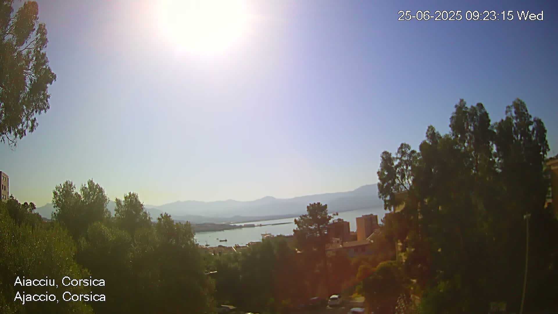 A sunny, clear day reveals a panoramic view of a coastal town nestled among green hills and a calm body of water with mountains in the background.