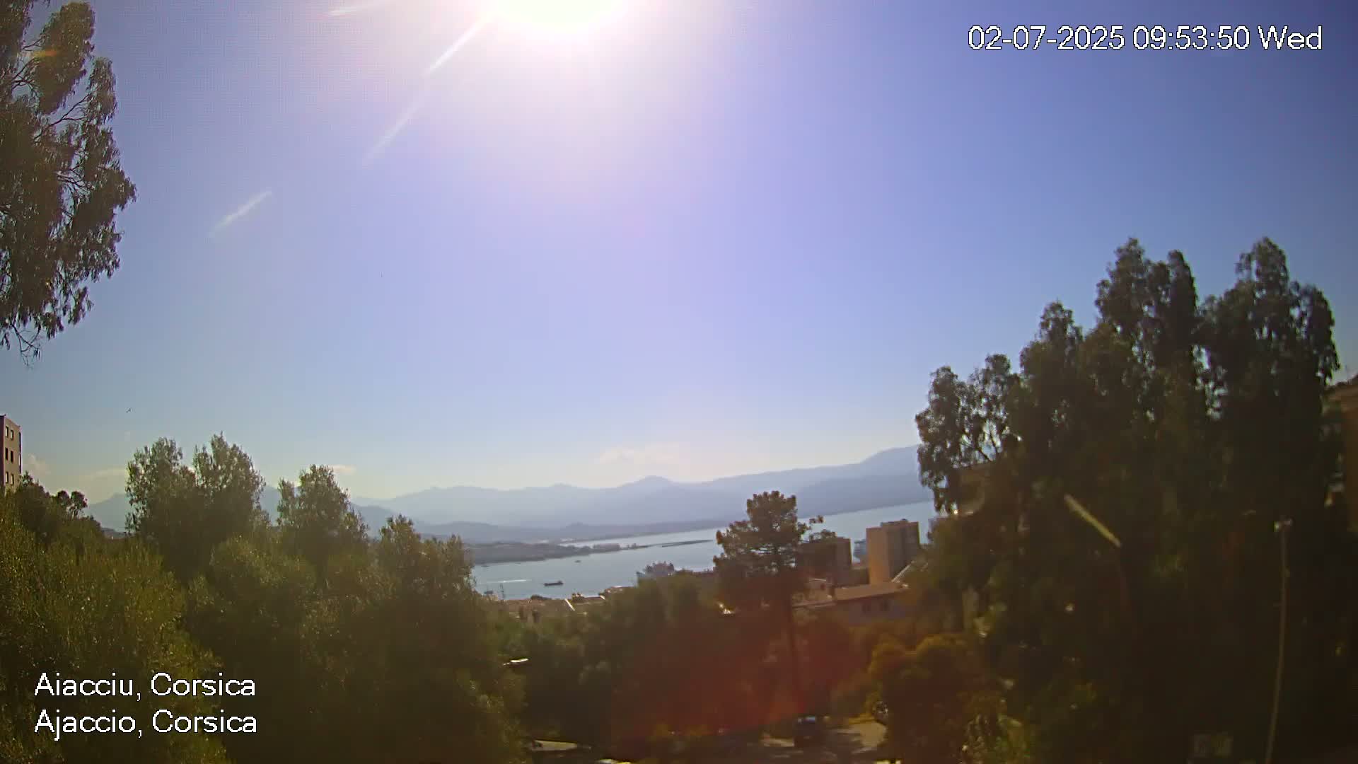 A sunny view overlooking a body of water, mountains in the distance, and a town nestled among green trees.