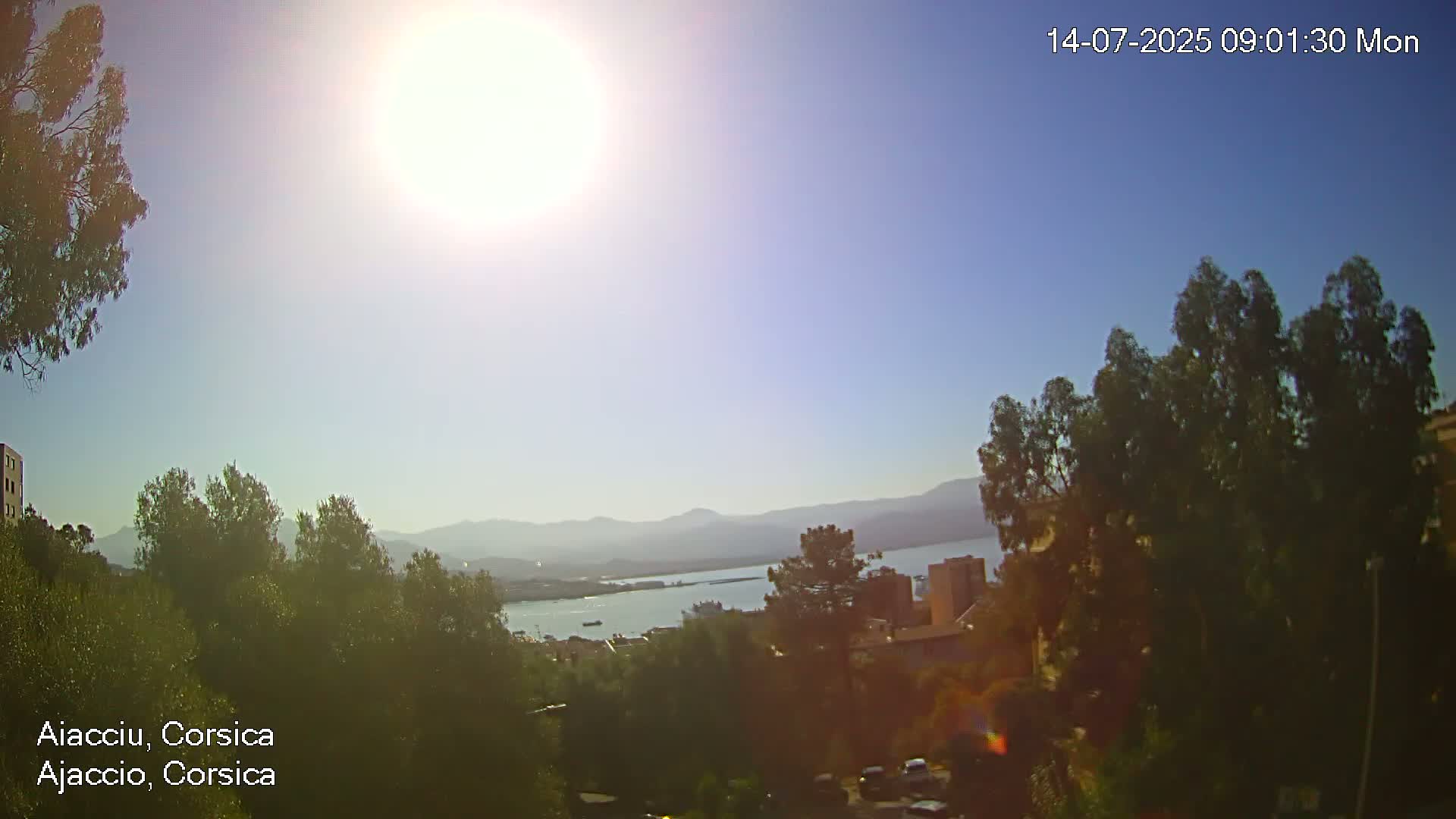 A bright, sunny day overlooks a body of water and distant mountains, with green trees and buildings in the foreground.