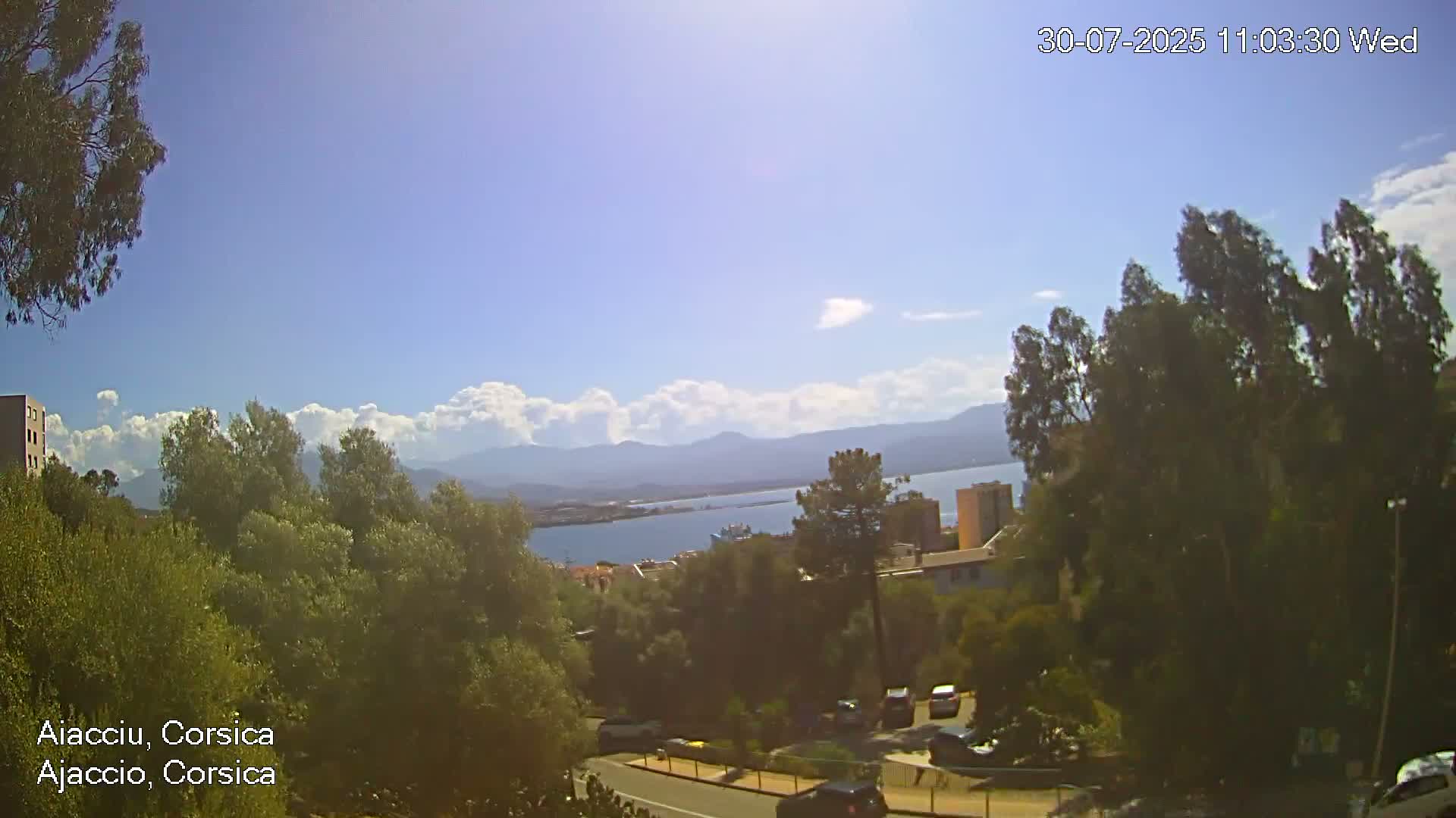 A sunny day with partly cloudy skies reveals a hilly coastal landscape with green vegetation, buildings, a body of water, and distant mountains.