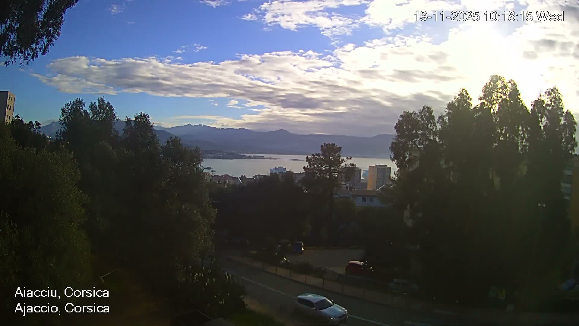 An elevated view reveals a coastal city nestled along a bay with distant mountains, a tree-lined road with cars in the foreground, all under a bright, partly cloudy sky.