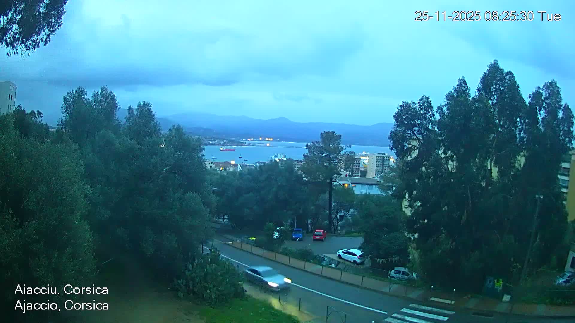 A rainy, overcast morning view captures a wet road with cars, lush green trees, coastal buildings, a bay dotted with boats, and distant mountains shrouded in mist.