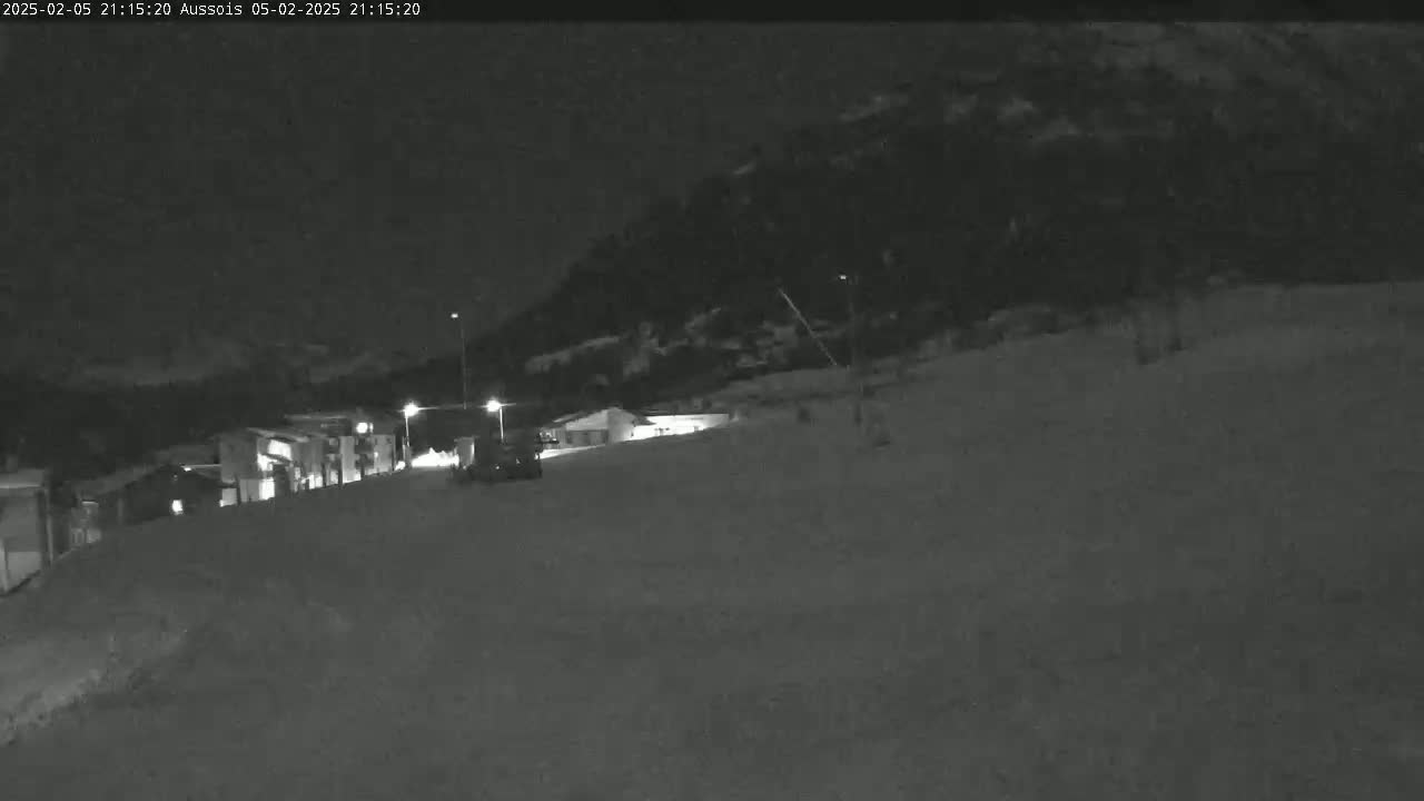 A nighttime, low-light view of a snow-covered area with buildings and a vehicle, illuminated by artificial lights against a dark mountain backdrop.