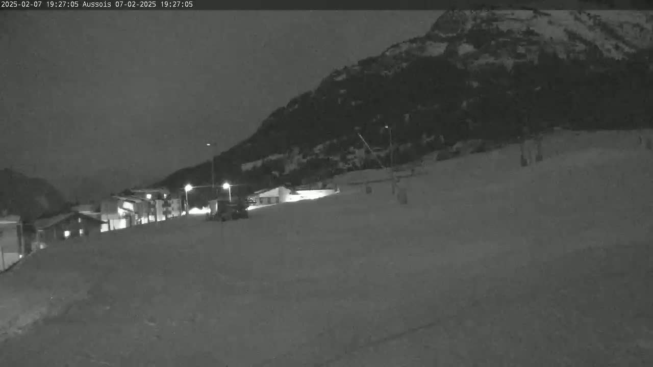 A nighttime, snow-covered scene shows a small village nestled at the base of a dark mountain.