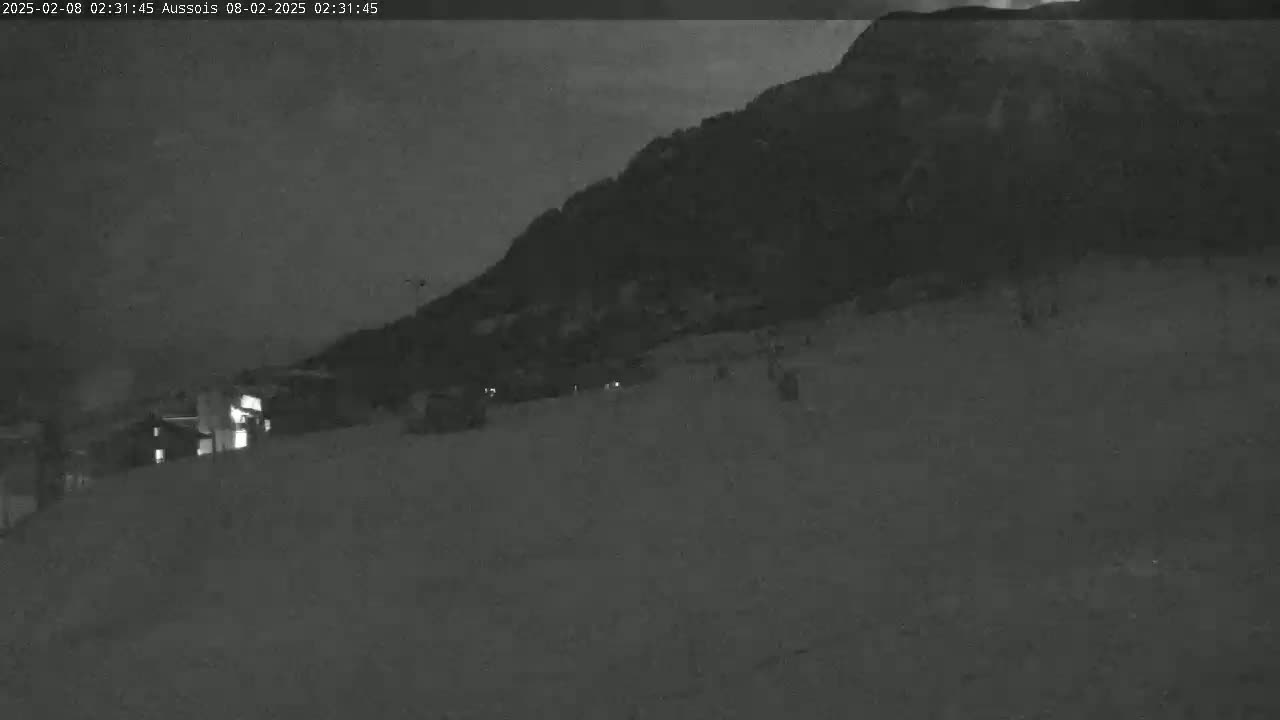 A dark, snowy landscape at night shows a large hill or mountain in the background with a few faint lights visible in the foreground.