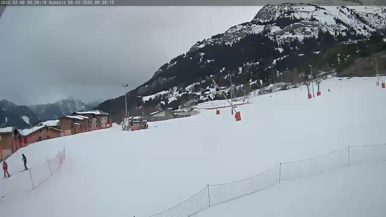 A snowy ski slope with a few skiers and a mountain in the background under an overcast sky.