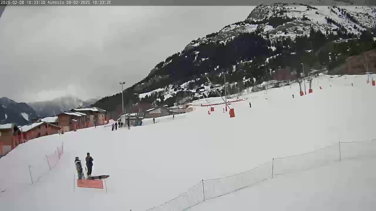 A snow-covered ski slope with several people skiing or snowboarding, nestled against a mountainside under an overcast sky.