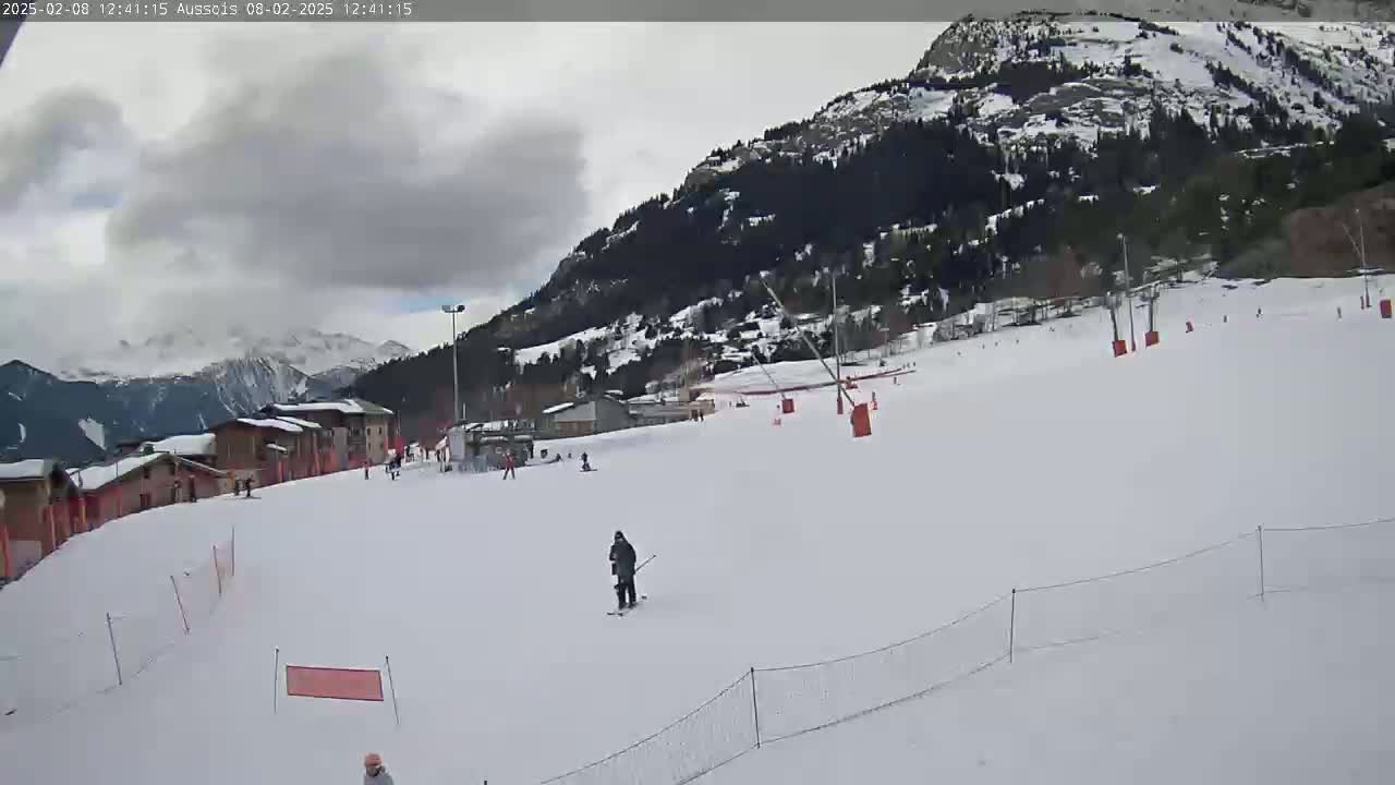 A mostly cloudy day on a snow-covered ski slope with a few skiers and a mountain in the background.
