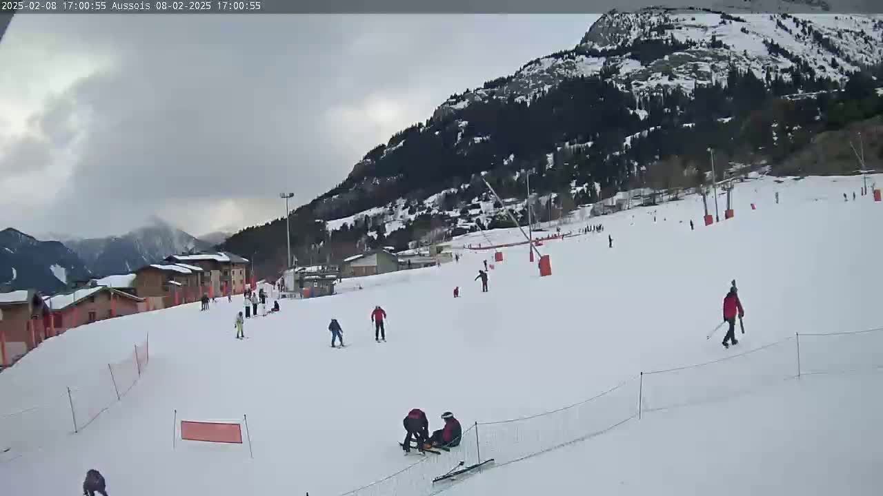 A snow-covered ski slope with several skiers and snowboarders, set against a backdrop of overcast skies and snow-dusted mountains.