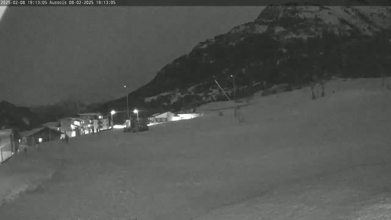 A nighttime, snow-covered scene shows a few buildings at the base of a dark mountain under a dark sky.