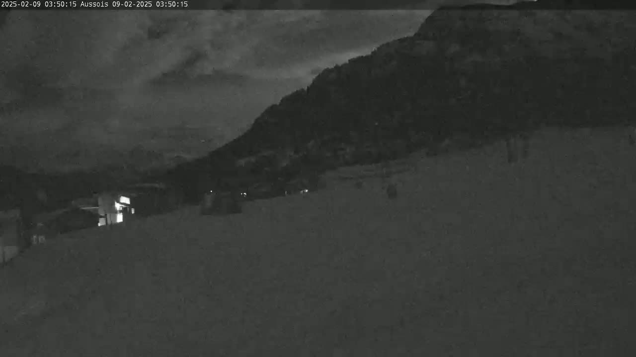 A nighttime, low-light view shows a snowy, flat area in front of a dark mountain under a cloudy sky.