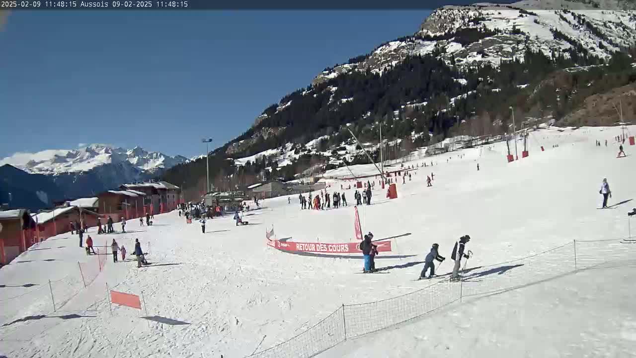 A sunny day on a snow-covered ski slope in a mountainous area, with several skiers and snowboarders present.
