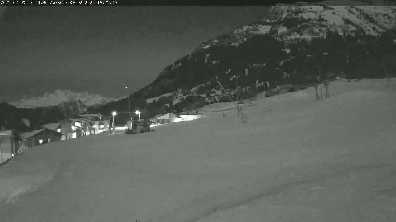 A snow-covered landscape at night, showing a village nestled at the base of a dark mountain under a clear sky.