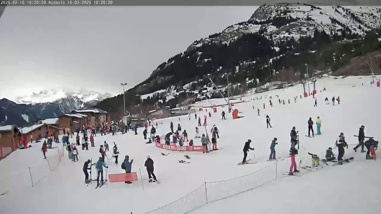 A snowy ski slope with numerous skiers and snowboarders, some buildings in the background, and overcast skies.