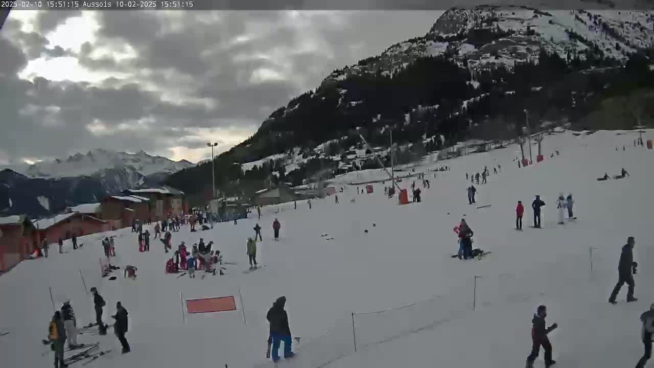 A snowy ski slope with numerous skiers and snowboarders is visible, under a mostly cloudy sky.