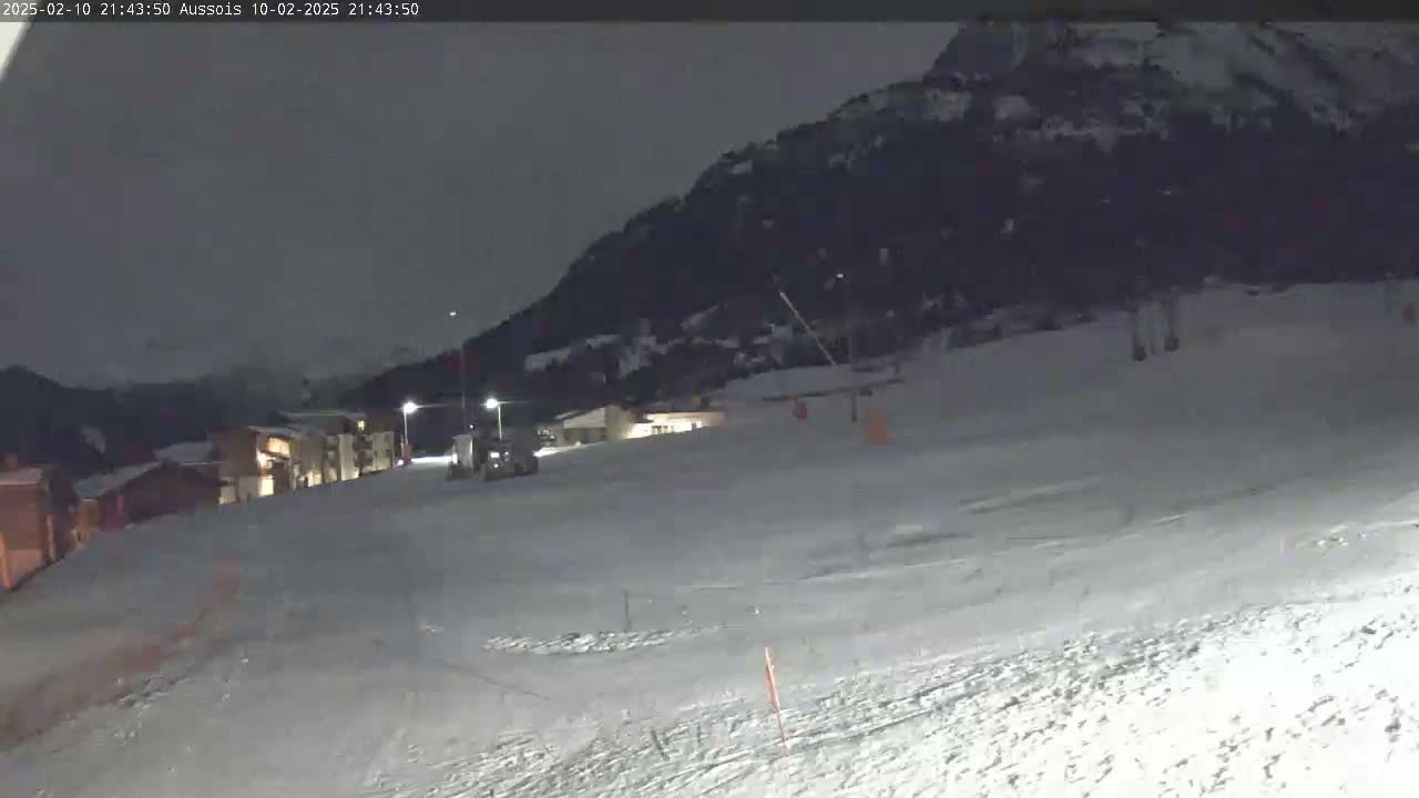 A snow-covered slope at night, illuminated by artificial lights, shows several buildings and a vehicle in the lower portion with a dark mountain in the background.