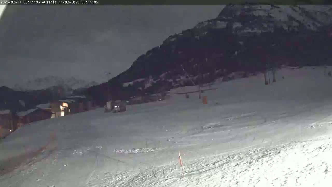 A snow-covered slope at night, with a few buildings and a mountain in the background under a dark, cloudy sky.