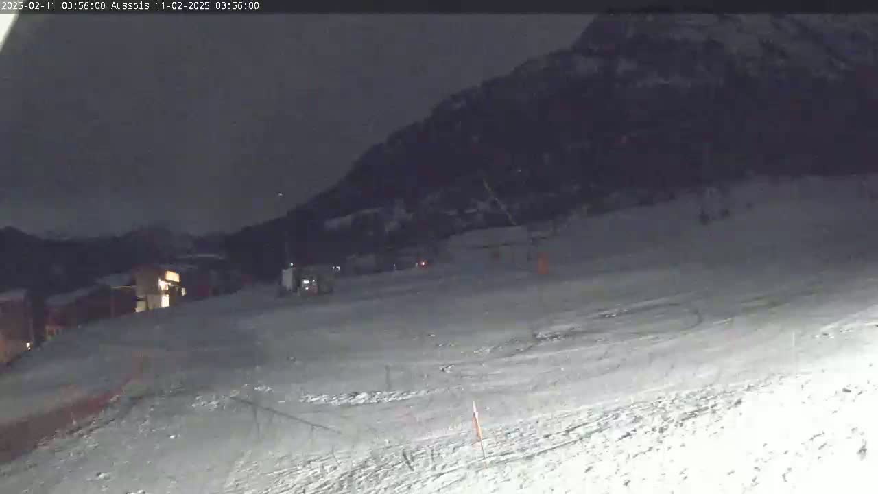 A snow-covered slope at night is illuminated, showing a few distant buildings and a dark mountain in the background.