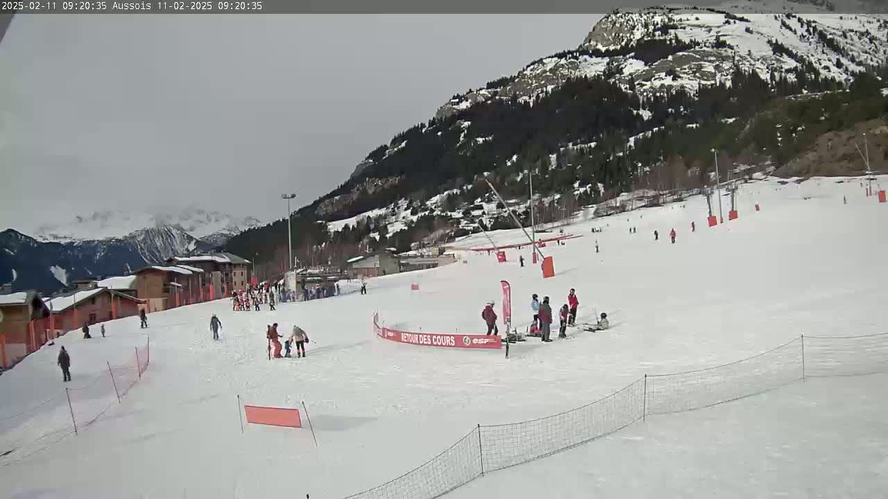 A snowy ski slope with several skiers and snowboarders, buildings, and a mountain in the background under an overcast sky.