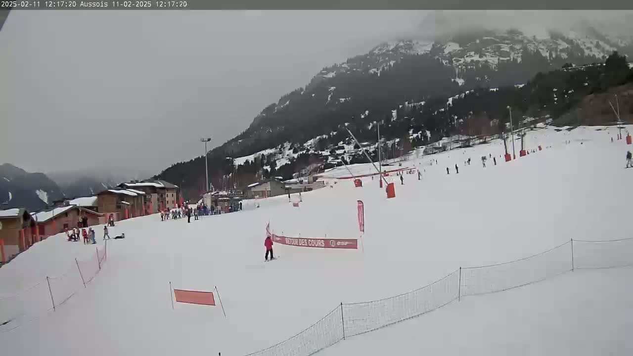 A snowy ski slope with several skiers and snowboarders, ski lifts, and buildings in the background under an overcast sky.