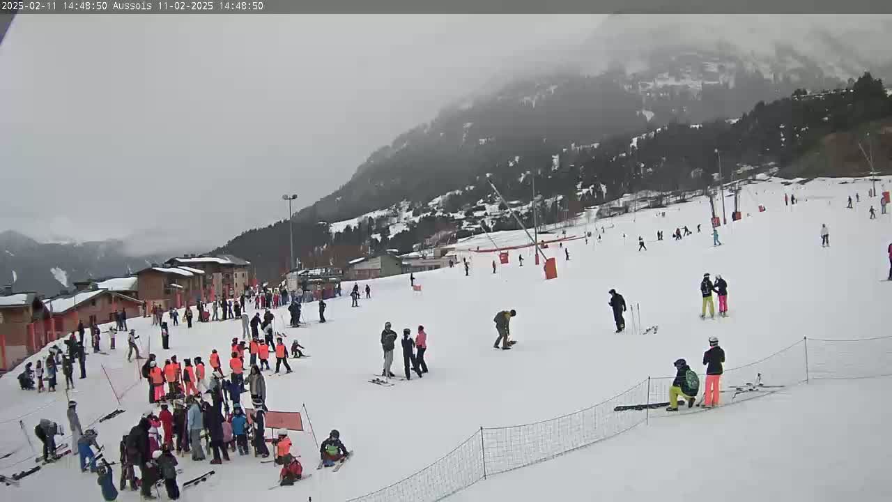 A snowy ski slope with numerous skiers and snowboarders, buildings, and a mountain in the background under overcast skies.