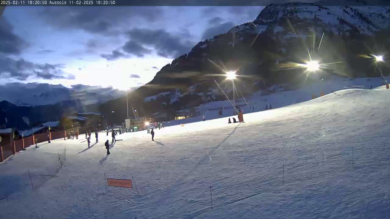 A snow-covered ski slope at dusk, illuminated by artificial lights, with a few skiers visible against a backdrop of mountains under a partly cloudy sky.