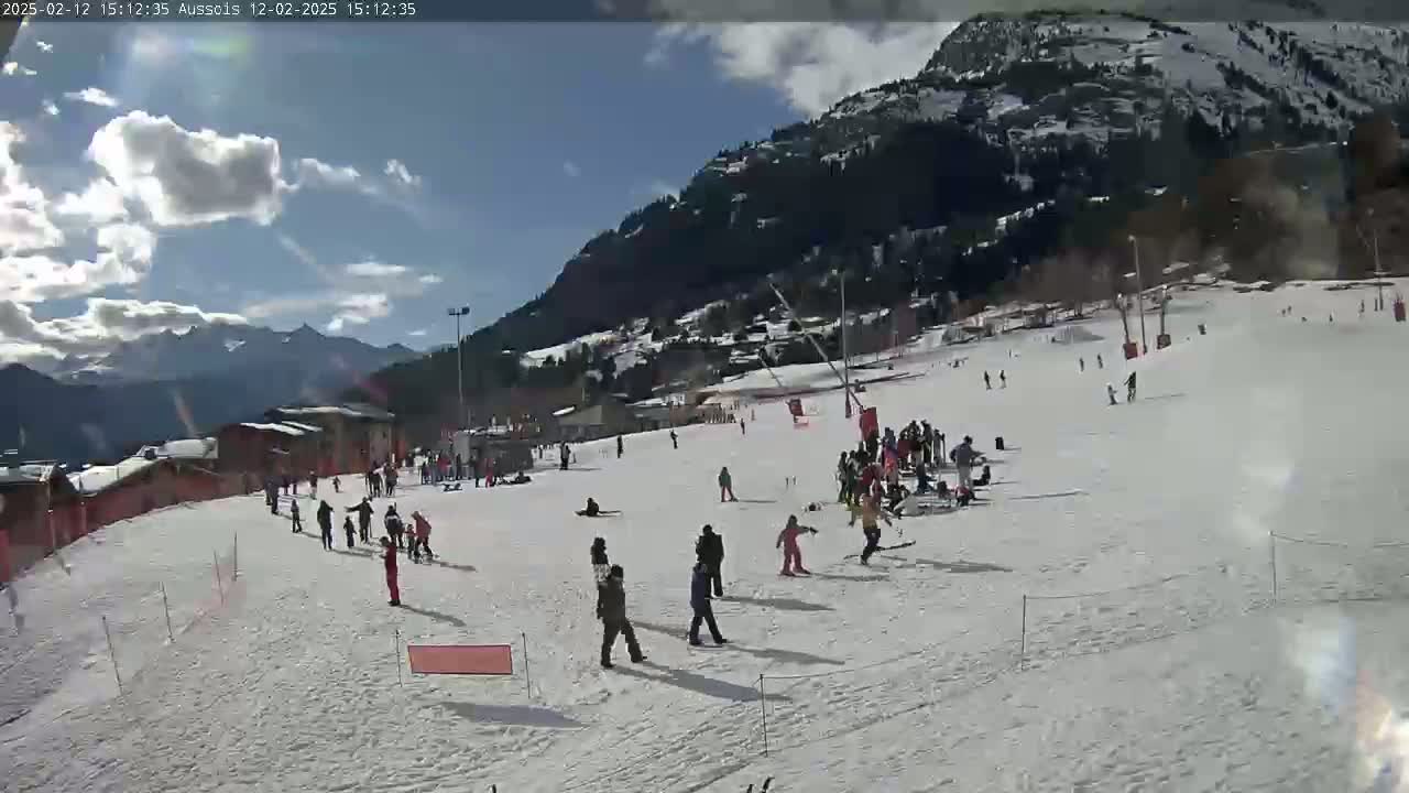 A snow-covered ski slope with several people skiing and snowboarding under a partly sunny sky.