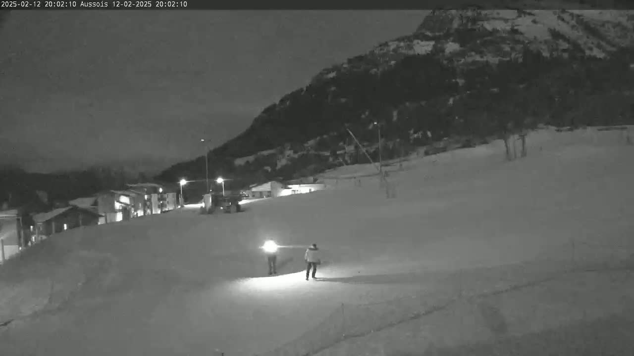 Two figures walk across a snow-covered slope at night under a dark sky, with buildings and a mountain in the background.