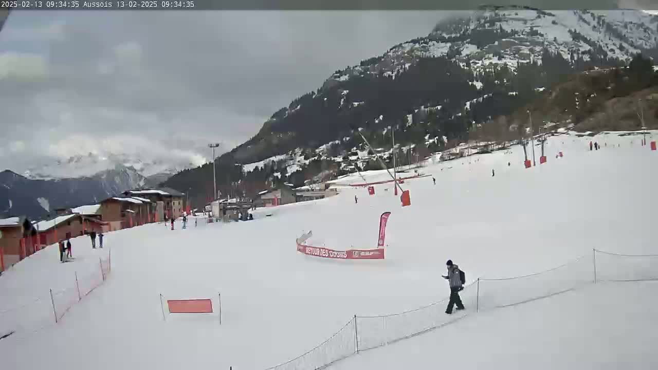 A snowy ski slope with several skiers and a person walking, nestled in a mountainous area under an overcast sky.