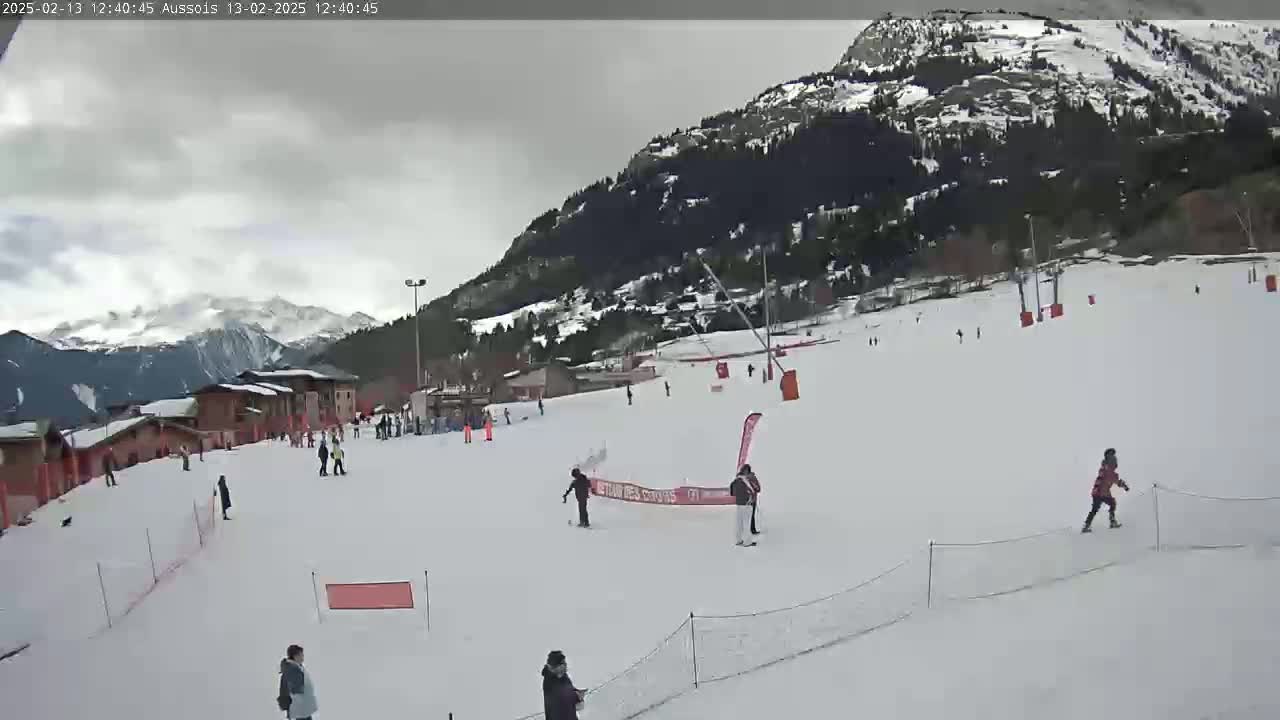 A snow-covered ski slope with several skiers and snowboarders, under an overcast sky, is nestled against a mountain range.