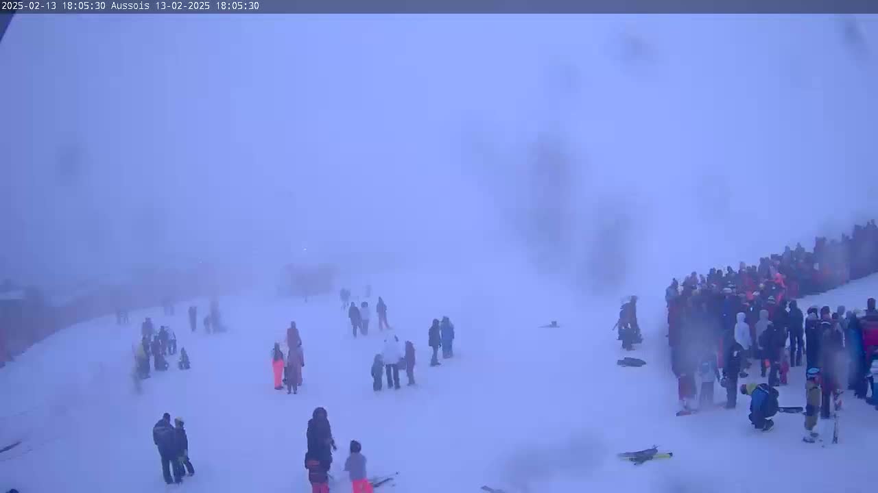 A snowy, foggy scene shows several small groups and a large crowd of people gathered on a wide, snow-covered slope.
