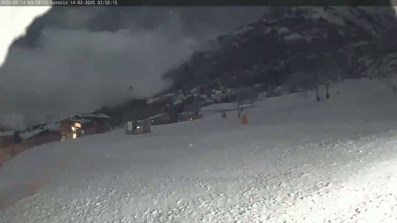 A snow-covered landscape at night is shown, with a mountain in the background under a heavy, overcast sky, and some buildings and what appears to be a ski lift visible in the foreground.