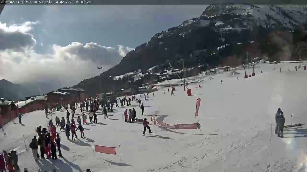 A snowy ski slope with numerous skiers and snowboarders, partially sunny weather, and a mountain backdrop.