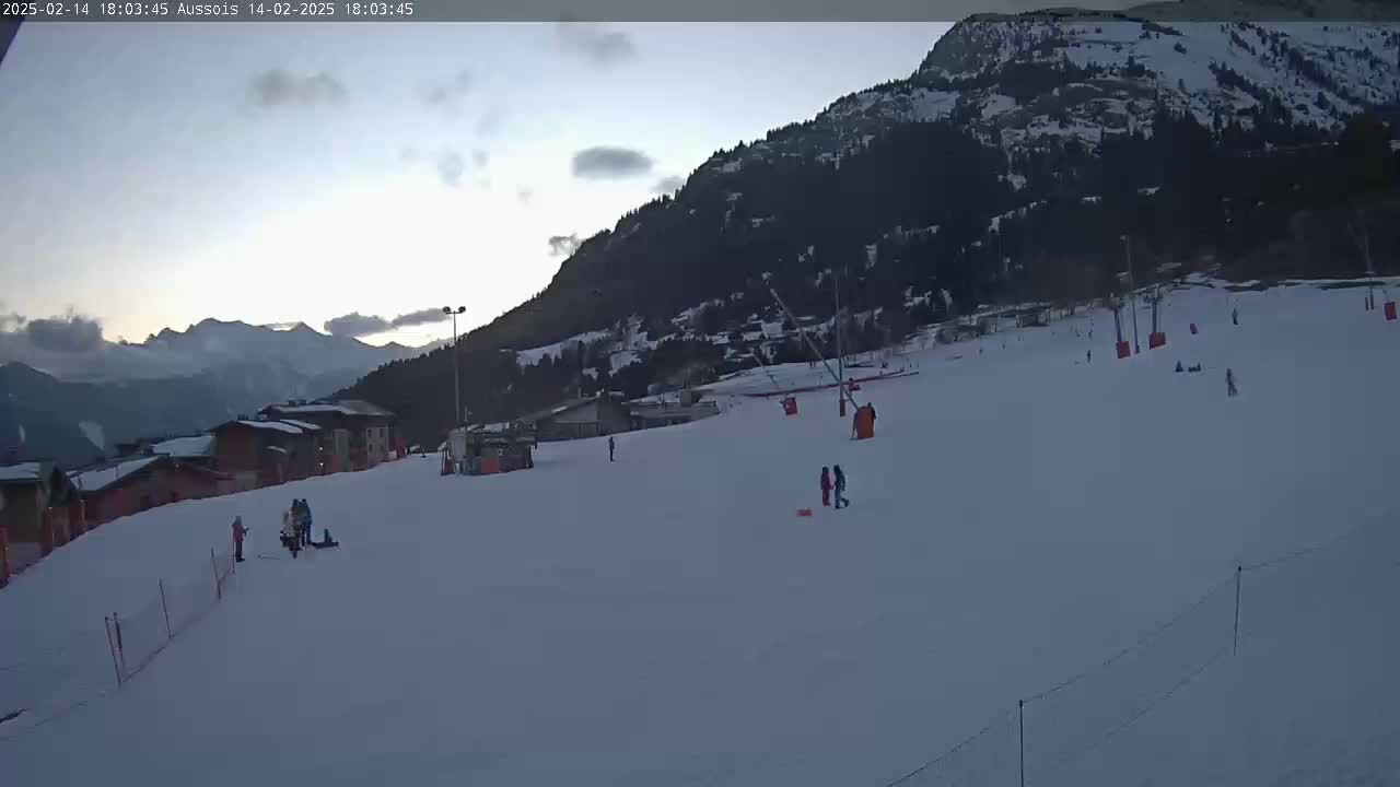 A snow-covered ski slope at dusk, with a few people scattered across it, nestled at the base of a dark mountain range under a cloudy sky.